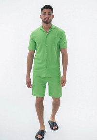 Chemise verte à manches courtes avec des boutons et une poche poitrine, assortie à un short. Le modèle porte des sandales noires à glissière. Tissu lisse et léger.