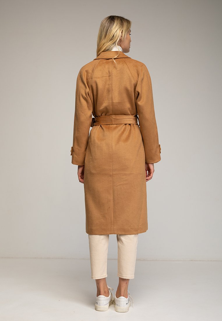 Cappotto lungo marrone con vita a cintura, realizzato in tessuto morbido. Presenta un collo a camicia e dettagli sui polsini, abbinato a pantaloni chiari e sneakers.