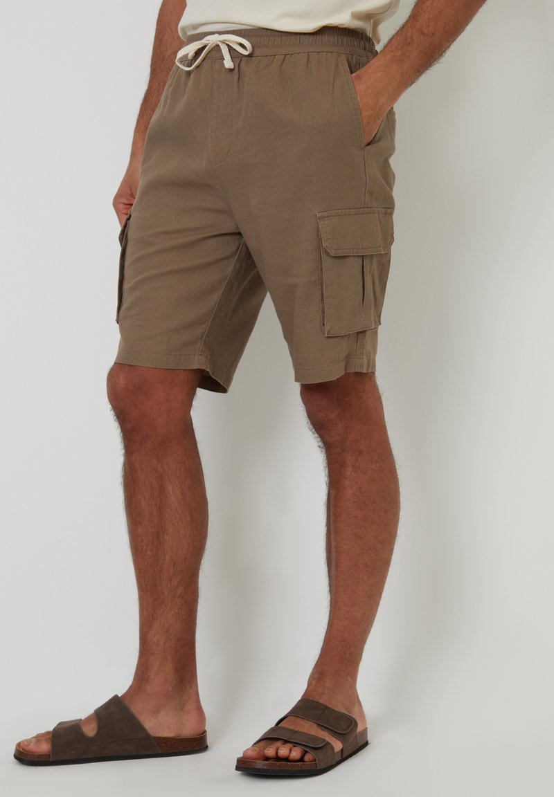 Khaki cargoshorts met een elastische tailleband en trekkoord, voorzien van zijzakken en een ontspannen pasvorm. Gecombineerd met bruine slippers.