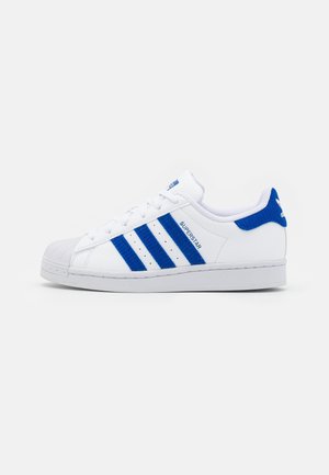 Hvid Adidas Superstar sneaker med blå striber, blå hælstrop, skal-tå og "Superstar" skrevet på siden.