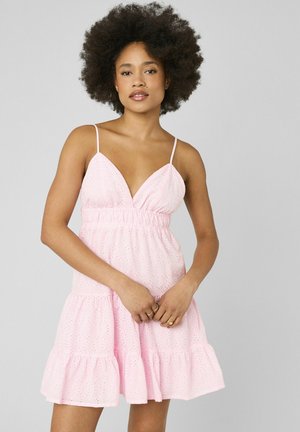 Femme aux cheveux bouclés portant une robe d'été rose sans manches en broderie anglaise avec un ourlet volanté, debout devant un fond uni gris clair.