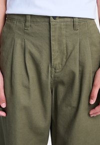 Pantaloni verde oliva realizzati in tessuto strutturato con vita alta, caratterizzati da pieghe frontali e chiusura con bottone.