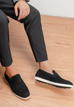 Personne assise les jambes croisées portant des chaussures noires à enfiler et un pantalon noir sur un sol en bois clair.