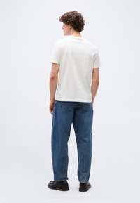 Νεαρός άνδρας με καστανά σγουρά μαλλιά στέκεται με την πλάτη, φορώντας λευκό κοντομάνικο t-shirt, μπλε τζιν και μαύρα παπούτσια σε απλό φόντο.