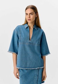 Haut en denim avec un décolleté en V, des manches courtes et des bords effilochés. Tissu bleu clair avec des coutures contrastées. Associé à une jupe assortie.