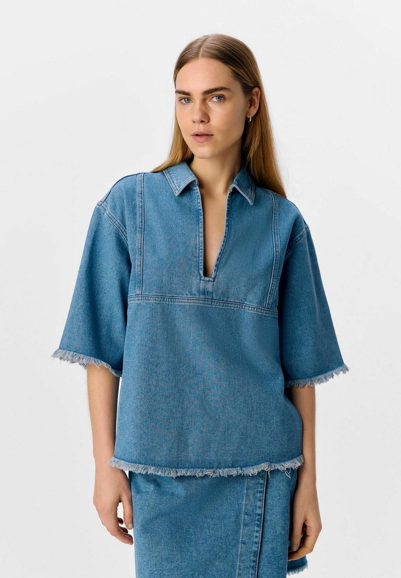 Haut en denim avec un décolleté en V, des manches courtes et des bords effilochés. Tissu bleu clair avec des coutures contrastées. Associé à une jupe assortie.