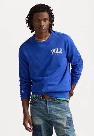 Sweatshirt bleu cobalt avec col rond et logo "Polo Ralph Lauren" blanc. Porté par-dessus une chemise superposée multicolore. Jean en denim avec des patchs.