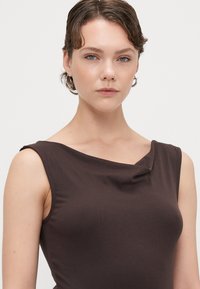 Smeđa majica bez rukava s cowl izrezom, izrađena od mekog materijala, ima prilagođeni siluetu i glatku teksturu. Suptilni detalji oko ramena.