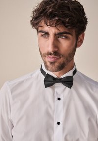 Bílá košile s černým motýlkem, s úzkým límečkem a třemi černými knoflíky. Hladká tkanina s jednoduchým designem a klasickou elegancí.