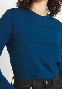 Long-sleeve teal cotton shirt with a crew neck. Features a smooth texture and fitted silhouette, paired with high-waisted black jeans.