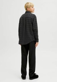 Melnā un baltā kvadrātveida flanelēna krekls, garām piedurknēm, izliekta apakšmala, apvienots ar melniem biksēm un melnām kurpēm, skatīts no aizmugures.