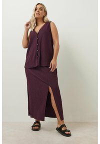 V-ringad, ärmlös topp och matchande kjol i djup lila, med ett strukturerat mönster och en sidoöppning. Svarta sandaler med nitar.