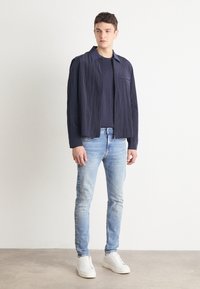 Chaqueta texturizada azul marino con cuello, sobre una camisa oscura, combinada con jeans de mezclilla azul claros y zapatillas blancas. Diseño simple y moderno.