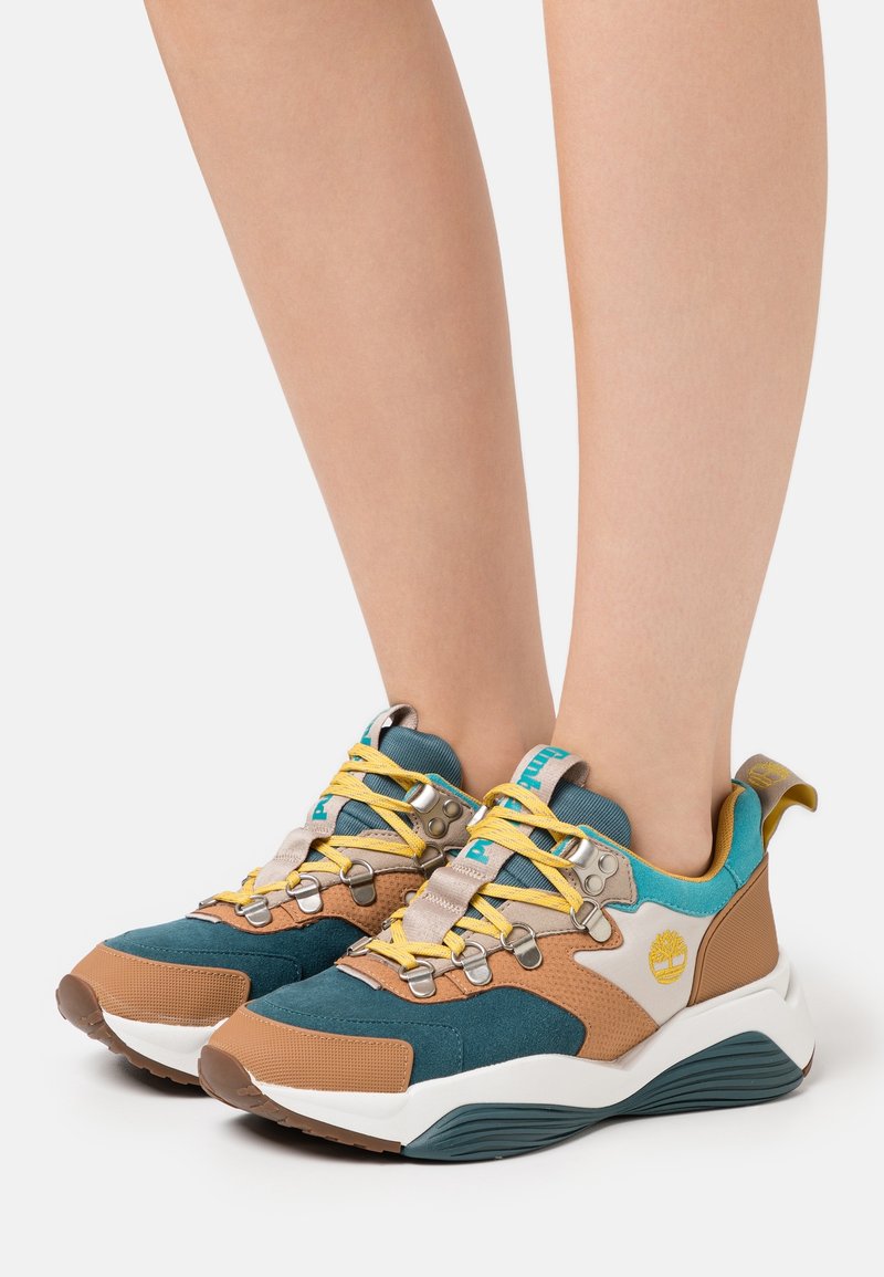 Jambes portant des baskets Timberland bleu sarcelle, marron et beige avec des lacets jaunes, des semelles intermédiaires blanches et des semelles robustes sur fond blanc.