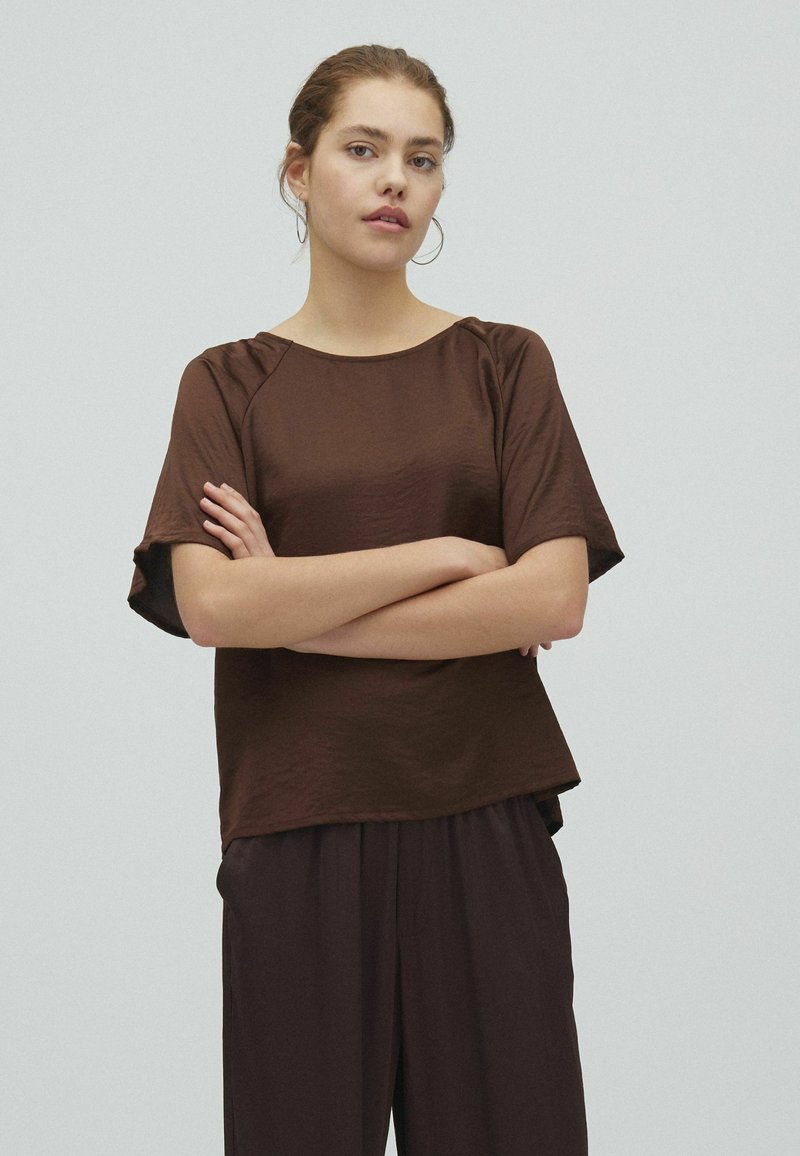 Jeune femme aux cheveux châtain clair attachés, portant des boucles d'oreilles créoles, un haut marron ample à manches courtes et un pantalon marron assorti, bras croisés.