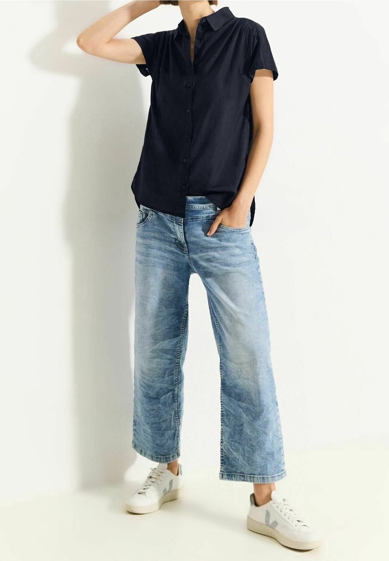 Personne portant une chemise noire à manches courtes boutonnée, un jean large bleu clair et des baskets blanches, posant avec une main dans la poche.