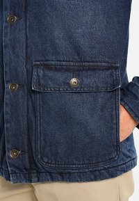 Tmavě modrá džínová bunda se zapínacím kapsou na knoflíky, kovovými knoflíky a detailem na manžetě. Textura je hladká s viditelným prošitím.