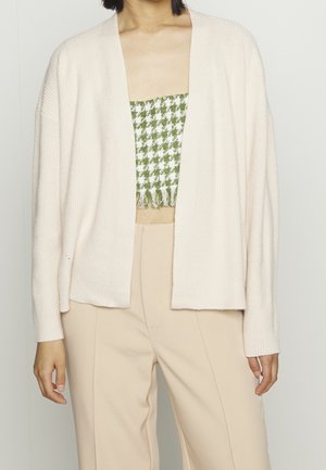 Vrouw die een crème open-front cardigan draagt, een groen-wit houndstooth crop top en beige hooggetailleerde broek, staand tegen een effen achtergrond.