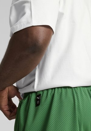 Bílá bavlněná tričko s krátkým rukávem je kombinováno se zelenými síťovanými šortkami s elastickým pasem a černou štítkovou značkou.