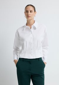 Witte, cropped knoopsluiting blouse met een klassieke kraag, een getailleerde pasvorm en lange mouwen; gecombineerd met donkergroene broeken. Gladde stofstructuur.