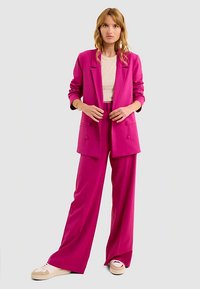 Costume fuchsia avec un blazer ajusté et un pantalon large, associé à un haut de couleur claire et des baskets neutres. Texture lisse, design moderne.
