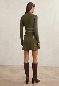Olīvu zaļš ribots mini kleita ar garām piedurknēm, ar baltām šuvju detaļām. Saskaņots ar ceļgalu augstiem brūniem ādas zābakiem.