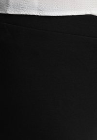 Fekete nyújtható anyag, sima textúrával, testhez álló fazonban. A felső éle fehér, kontrasztot teremtve a feketével.