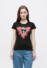 Čierne bavlnené tričko s priliehavým strihom, na ktorom je červený kvetinový trojuholníkový motív a text "GUESS?" v bielej a čiernej farbe v rámci ohraničenia.