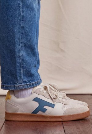 Jambes en jean bleu et baskets blanches avec un logo "F" bleu et un languette talon jaune, debout sur un sol en bois devant un fond beige.