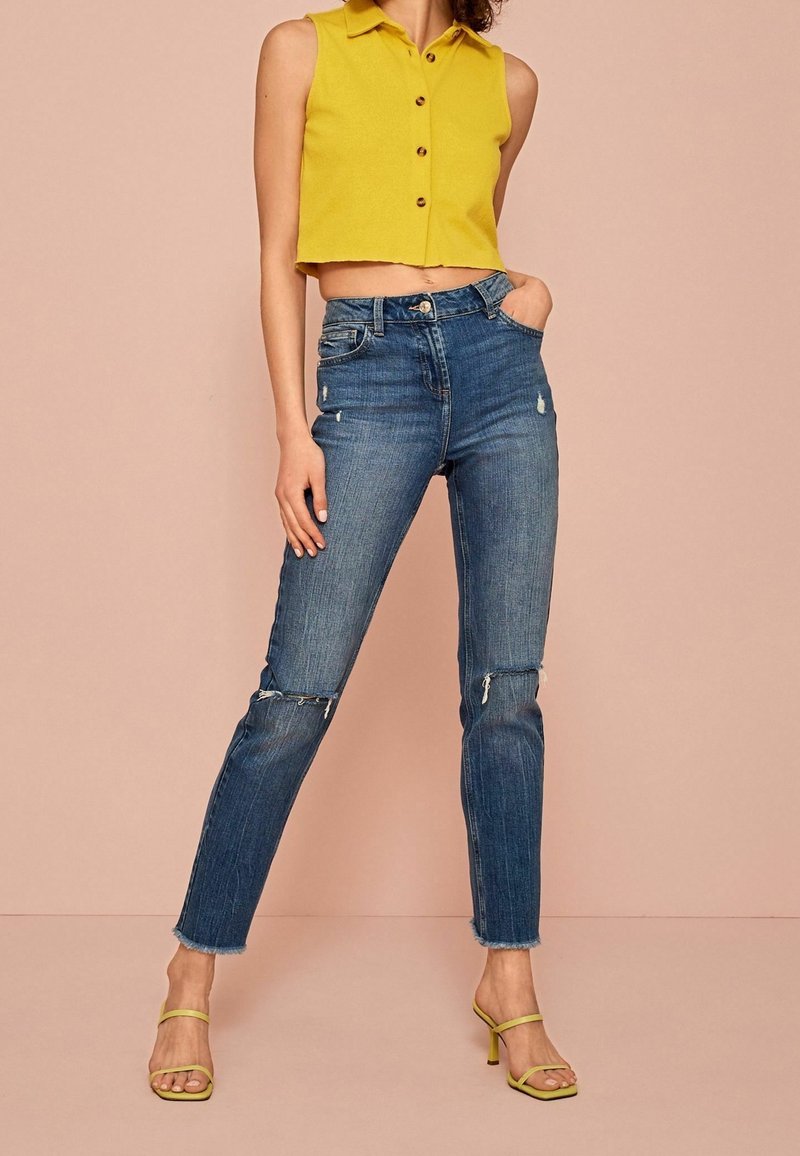 Una modelo lleva una blusa amarilla sin mangas y jeans azules desgastados, de pie sobre un fondo rosa claro con tacones amarillos con tiras.
