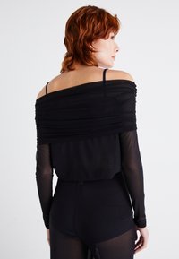 Μαύρο τοπ χωρίς ώμους με ρυάκια, διαφανή μακριά μανίκια και εφαρμοστή σιλουέτα. Το υλικό φαίνεται απαλό και ελαφρύ.