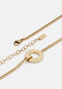 Gouden ketting met een cirkelvormige hanger met de gravure "BOSS", een kreeftsluiting, en afwisselende schakels van verschillende maten en texturen.