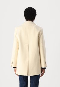 Cappotto di lana color crema con un design strutturato, ampi revers e una fessura singola sul retro. Presenta maniche con bottoni e una silhouette slanciata.