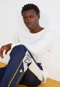 Camicia bianca a maniche lunghe e pantaloni sportivi blu con accenti gialli e logo. Modello seduto contro un muro bianco, che mette in risalto i dettagli dell'abbigliamento.