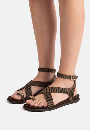 Pies con sandalias planas con tiras a rayas marrones y negras, hebilla en el tobillo y anillos dorados en los dedos, sobre un fondo neutro.