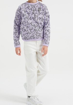 Enfant portant un pantalon blanc, des baskets blanches et un t-shirt à manches longues lavande avec un motif moucheté noir et un patch tigre.