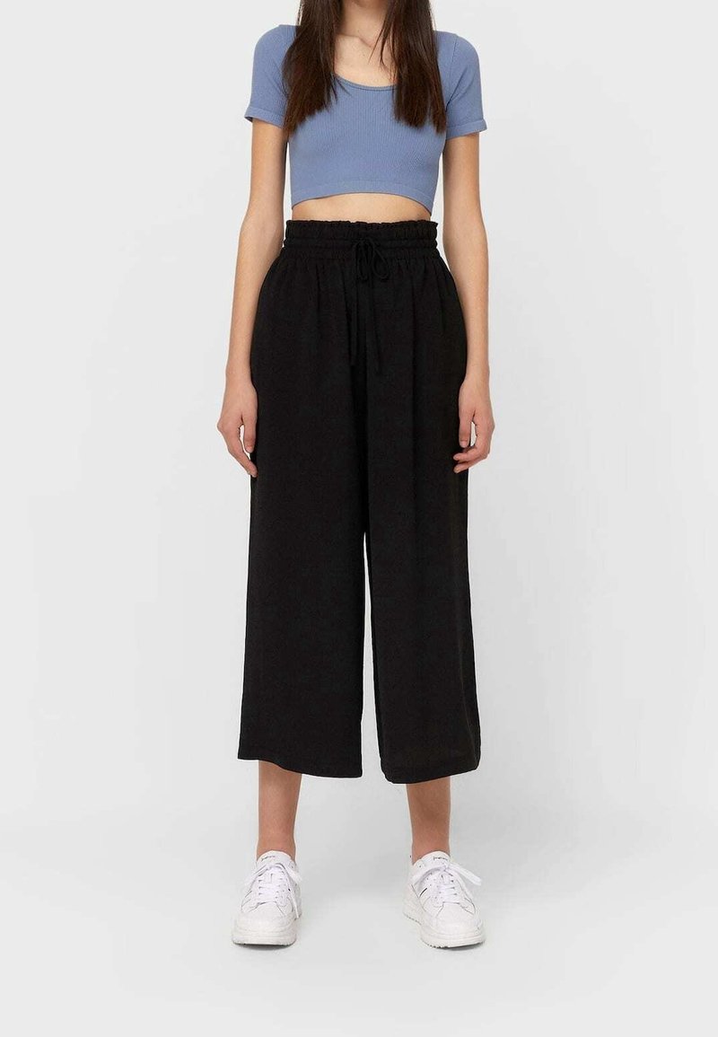 Femme portant un haut court bleu à manches courtes, un pantalon noir large et court à taille élastique avec cordon, et des baskets blanches, debout sur un sol gris.
