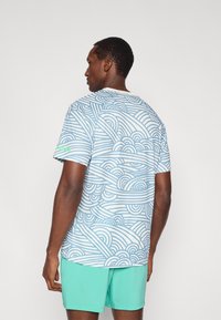 Κοντομάνικο t-shirt σε ανοιχτό μπλε με λευκό σχέδιο κυμάτων, κατασκευασμένο από απαλό ύφασμα. Διαθέτει στρογγυλή λαιμόκοψη και αντίθετο πράσινο λογότυπο στο μανίκι.