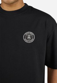 Svart bomullst-skjorte med et sirkulært brodert logo med teksten "Everyday Ambition" og "University of Pegador" i hvitt. Korte ermer.