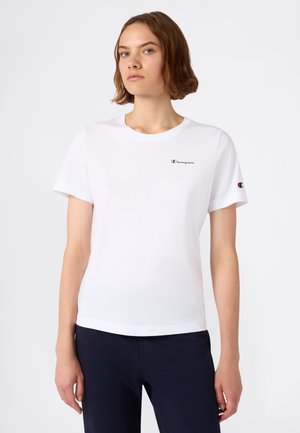 Giovane donna che indossa una t-shirt bianca Champion con il logo sul petto e sulla manica, abbinata a pantaloni scuri, in piedi davanti a uno sfondo semplice.