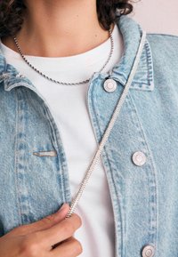 Person iført en sølv kæde halskæde, hvid skjorte og en lys blå denimjakke, der holder et rhinestone-rem diagonalt over brystet.