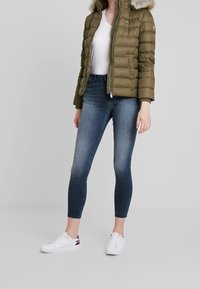 Olivgrön puffjacka med en huva klädd i päls, dragkedja fram, ribbade ärmar, kombinerad med mörkblå tajta jeans och vita sneakers.