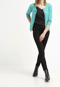 Turkos kofta, svart topp med rundad halsringning, svarta jeans och svarta ankelboots. Koftan har en knappstängning och långa ärmar.