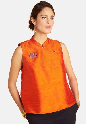Frau trägt ärmelloses, leuchtend orangefarbenes Oberteil mit Stehkragen und lila Elefantenbrosche, Hände in den Taschen schwarzer Hosen, goldener Schmuck.