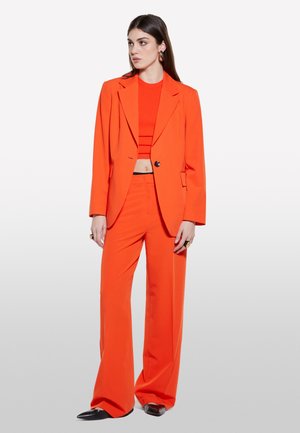 Completo arancione composto da una giacca corta e pantaloni a gamba larga. Il materiale sembra liscio, con un colore audace e dettagli minimi.