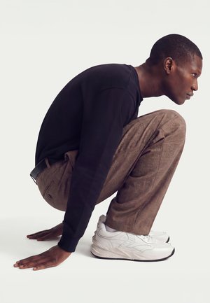 Mann in braunen karierten Hosen, schwarzem Langarmshirt und weißen Turnschuhen hockt mit den Händen auf dem Boden, nach rechts blickend vor einem einfarbigen Hintergrund.