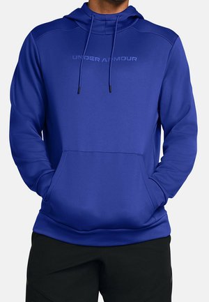 Mand iført en blå Under Armour hættetrøje med frontlomme og snore, hænderne i lommerne, kombineret med sorte bukser.