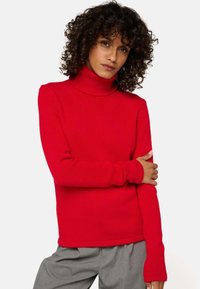 Personne avec des cheveux bouclés portant un pull col roulé rouge vif et un pantalon gris, posant avec un bras croisé sur l'autre.