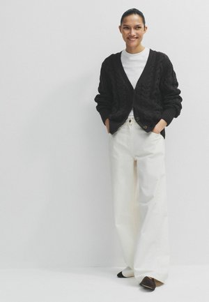 Mosolygó nő fekete kábelmintás kardigánt visel fehér ing fölött, széles fehér nadrágot és fekete hegyes cipőt, egyszerű világos háttér előtt áll.