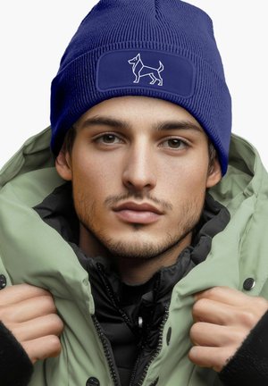 Navy knit beanie with a dog graphic and a patch. Worn with a green puffy jacket, showcasing a textured ribbed design.